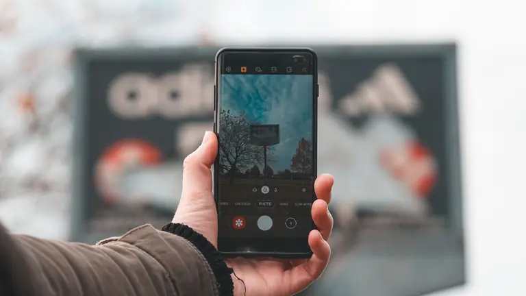 A hand holds a smartphone taking an autumn photo with trees in the background.
