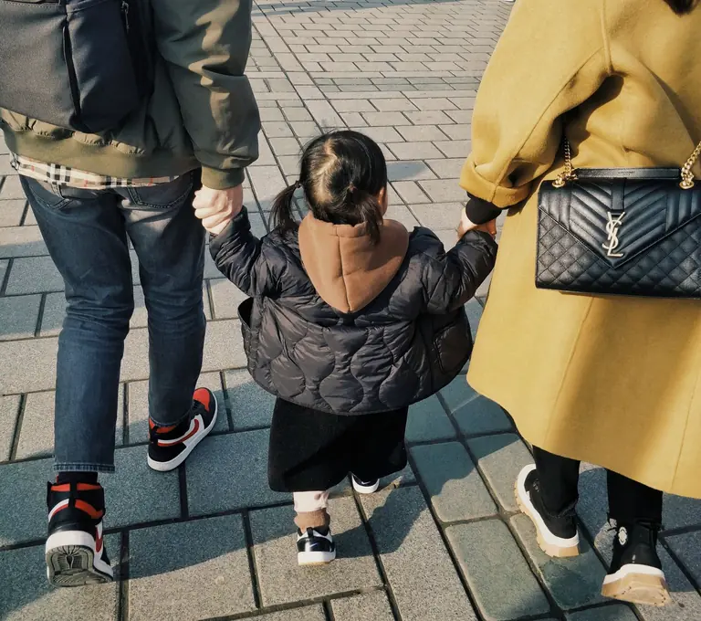 A family of three holding hands while walking on a city sidewalk in casual attire.