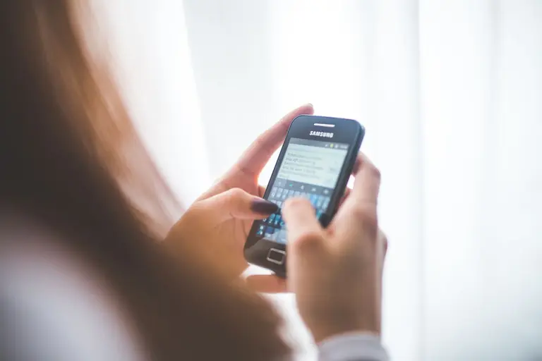 Close-up of a young woman typing on a Samsung smartphone at home. Bright indoor lighting.