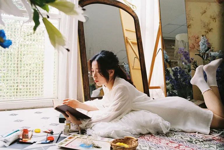 Asian woman reading a book indoors surrounded by art supplies and flowers.