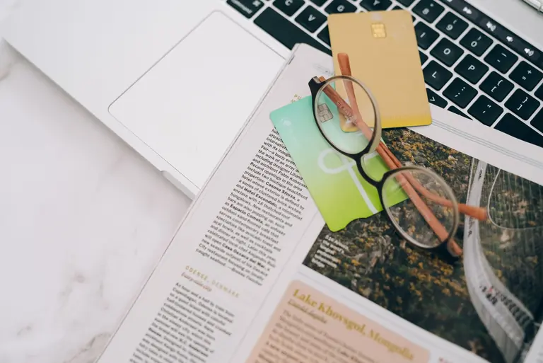 Eyeglasses resting on a magazine beside a laptop and credit cards, indoors.