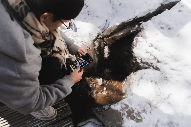 A person examines a smartphone close to a snow-covered stream.