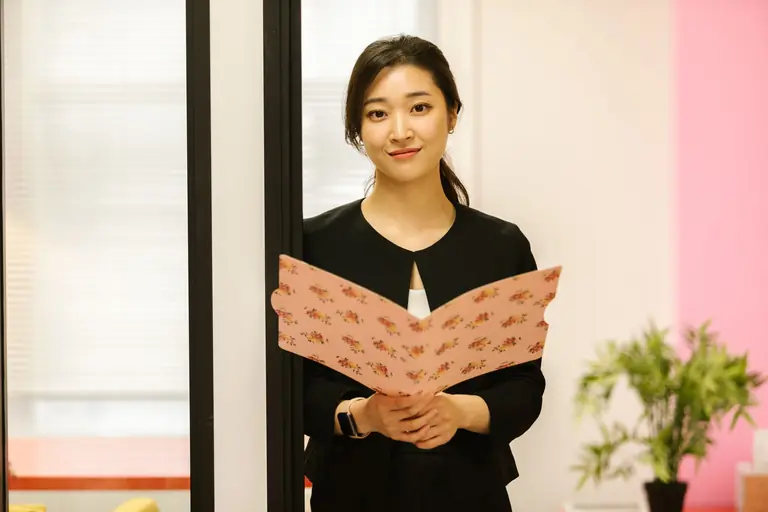 Confident woman in office attire holding a floral folder, standing in a modern workplace.