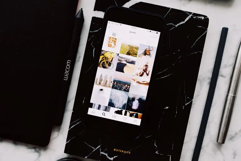 Top view of black notebook with cellphone above with pictures on screen and two pens aside located on marble table
