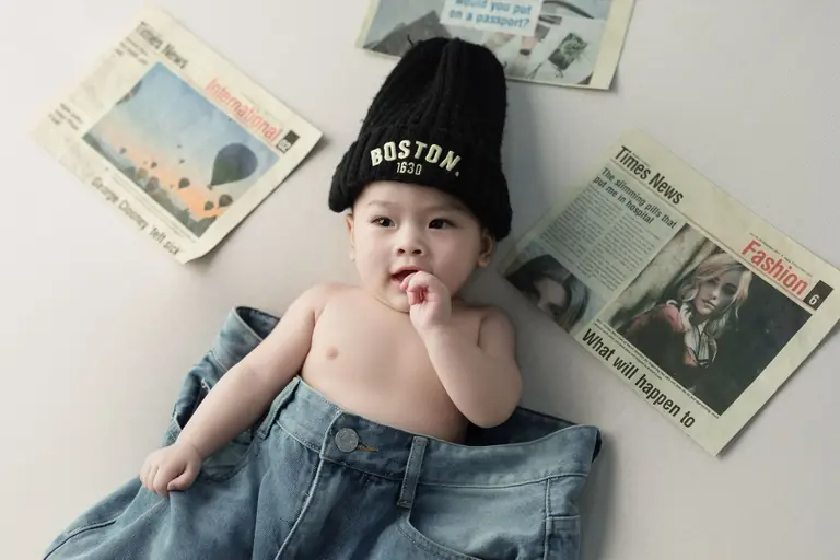 Adorable baby wearing jeans and beanie, surrounded by newspapers for a playful, modern look.