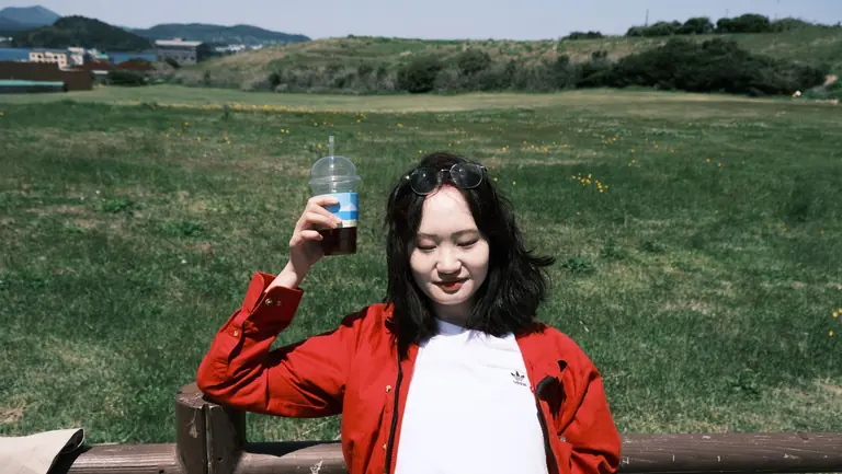 A young woman enjoys a sunny day outdoors, holding a drink and wearing sunglasses.