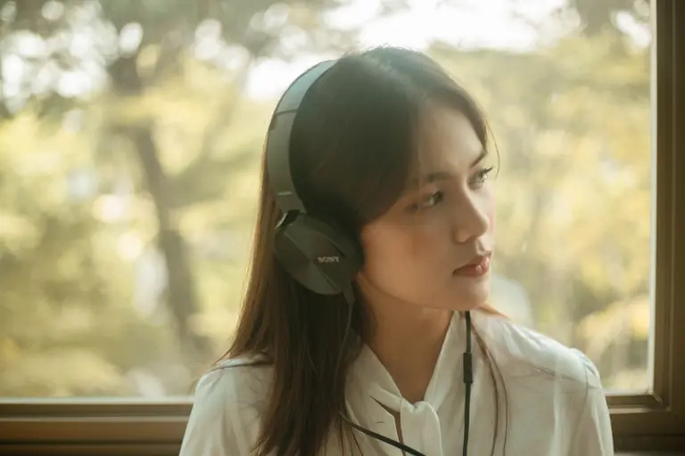 Portrait of a young woman with headphones enjoying music by a window with a serene expression.