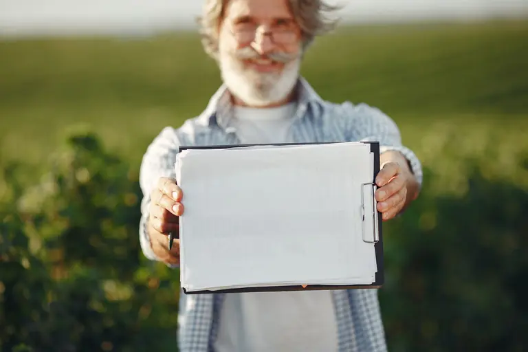 Bearded senior man with glasses holding a clipboard in a green field on a sunny day.