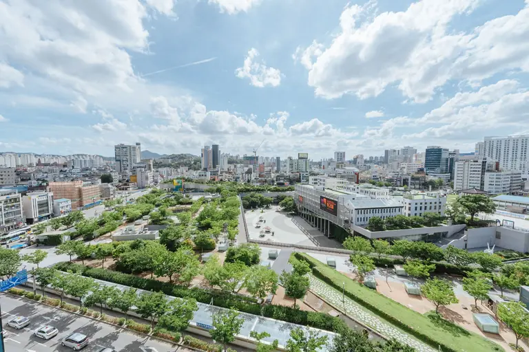 A vibrant cityscape featuring a lush urban park surrounded by modern buildings under a partly cloudy sky.